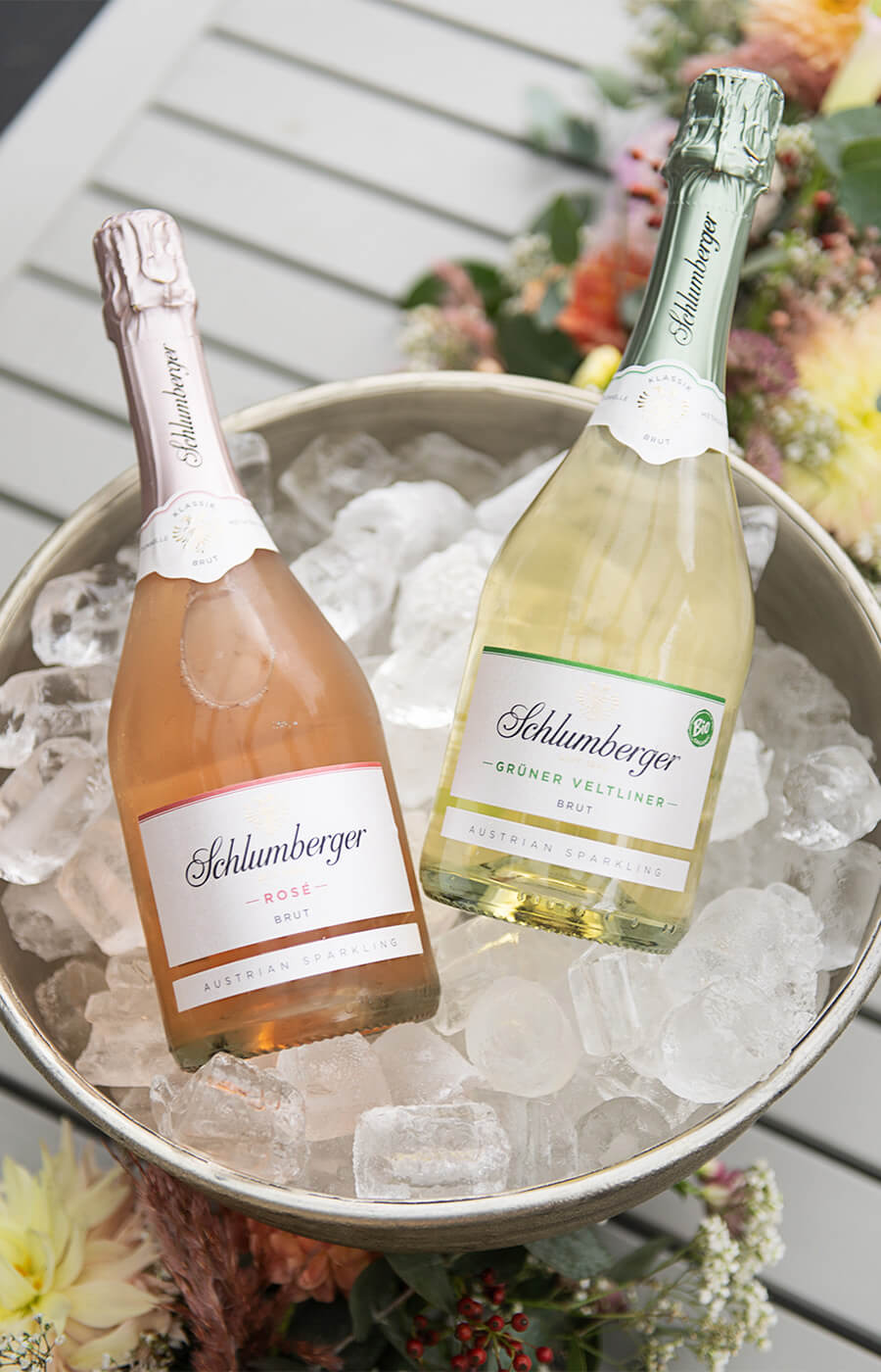 Zwei Flaschen Schlumberger-Sekt – Rosé Brut und Grüner Veltliner Brut – in einer Schale mit Eis, umgeben von Blumenarrangements.